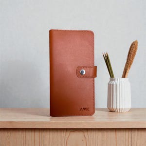 May include: A vertical, rectangular, tan leather wallet featuring a snap-button closure tab on the right side. The bottom right corner is embossed with the letters A and K separated by a small heart symbol. The leather has a smooth, matte finish.