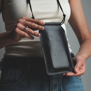 May include: A black rectangular leather phone pouch with a smooth finish and stitched edges. The pouch features a thin black cord strap attached to the top, allowing it to be worn around the neck or shoulder for carrying a mobile device.
