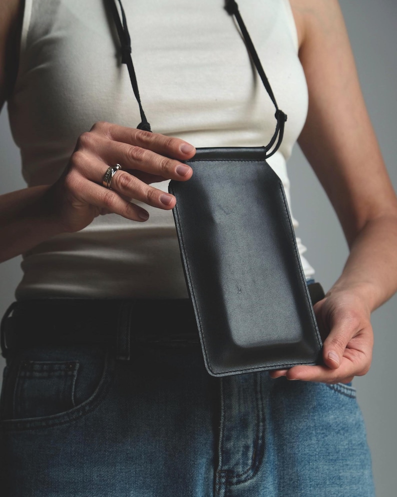 May include: A black rectangular leather phone pouch with a smooth finish and stitched edges. The pouch features a thin black cord strap attached to the top, allowing it to be worn around the neck or shoulder for carrying a mobile device.