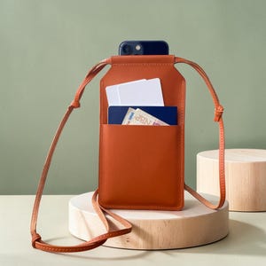 May include: A rectangular, burnt-orange leather crossbody phone pouch featuring a long, thin matching strap. The pouch has an open front pocket containing two white rectangular cards and a blue passport with visible text and graphics.