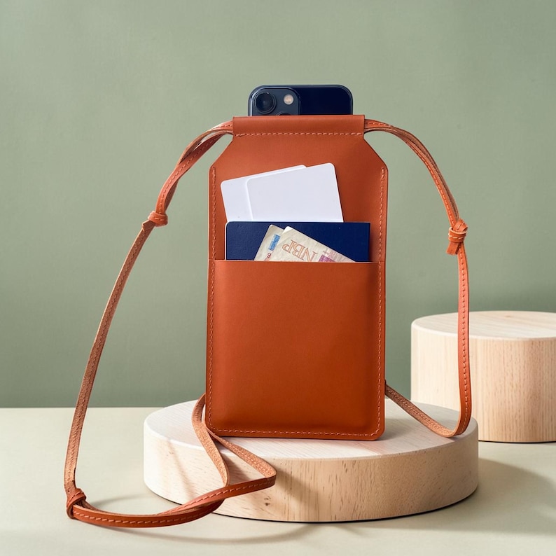 May include: A rectangular, burnt-orange leather crossbody phone pouch featuring a long, thin matching strap. The pouch has an open front pocket containing two white rectangular cards and a blue passport with visible text and graphics.