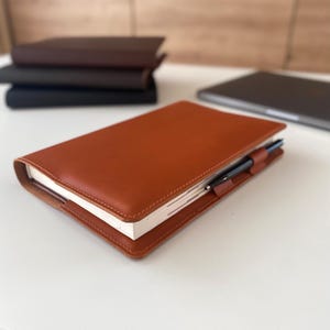 Könnte beinhalten: Ein Notizbuchumschlag aus hellbraunem Leder mit sichtbaren Nähten an den Kanten und einer integrierten Stiftschlaufe, in der ein schwarzer Stift steckt. Das Notizbuch liegt geschlossen auf einer weißen Oberfläche, im Hintergrund sind weitere Notizbücher gestapelt.