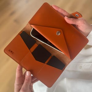 Op de afbeelding: Een bruine leren portemonnee met drukknoopsluiting, voorzien van een telefoonvak, meerdere kaartsleuven en de in reliëf aangebrachte letters A en K gescheiden door een hartje. De binnenzijde is gevoerd met bijpassend leer en bevat zwarte kaarten.