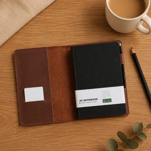 Op de afbeelding: Een donkerbruine leren notitieboekhoes met een zwart A5-notitieboekje voorzien van een witte papieren band met de tekst A5 NOTEBOOK. De hoes heeft een klein binnenvak en een penlus met een zwart potlood. Er staat een witte mok koffie bij.