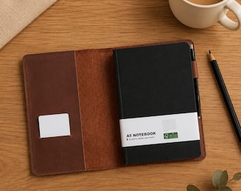 Puede incluir: Una funda de cuero marrón oscuro para cuaderno que contiene un cuaderno A5 negro con una banda de papel blanca etiquetada como A5 NOTEBOOK. La funda incluye un pequeño bolsillo interior y un portalápices con un lápiz negro. Una taza de café.