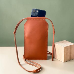 May include: A smooth, cognac-colored leather phone pouch with a long, thin matching shoulder strap. The rectangular pouch features clean, stitched edges and holds a dark blue smartphone that is partially visible at the top opening.