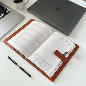 Può includere: Un'agenda aperta in pelle color cuoio con chiusura magnetica, dotata di pagine bianche con programmi giornalieri stampati, un calendario e il numero 2 sulla pagina destra. Una penna a sfera nera è appoggiata accanto all'agenda su una superficie bianca.
