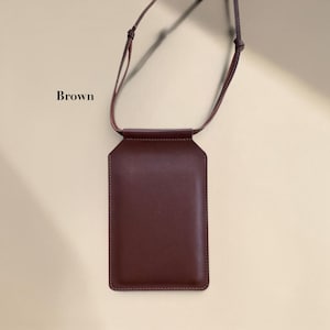 May include: A brown vertical rectangular pouch with a smooth finish and a thin matching shoulder strap. The pouch features a flap closure at the top and is labeled with the word Brown to the left of the strap attachment point.