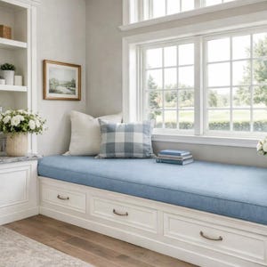 May include: A built-in window seat with a light blue cushion and white storage drawers. Two decorative pillows, one white and one blue plaid, rest against the window. A framed landscape painting hangs on the wall, and a stack of books sits on the seat.