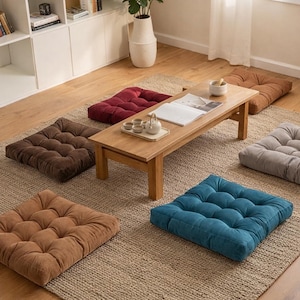 Puede incluir: Una mesa baja de madera rodeada de siete cojines cuadrados para el suelo en varios colores: marrón, rojo, beige, gris y verde azulado. Los cojines tienen un diseño acolchado. La mesa tiene una bandeja con tazas de té y una tetera.