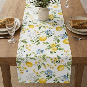 Può includere: Runner da tavolo bianco con un motivo a limoni e fiori. Il design presenta limoni gialli, fiori blu e foglie verdi. Il runner è posto su un tavolo di legno.