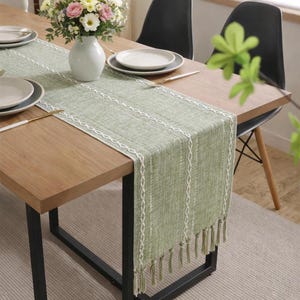 May include: A sage green table runner with white decorative trim and tassels. The runner is placed on a wooden table, with a vase of flowers and place settings visible. The table has black metal legs.