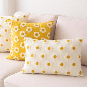 Op de afbeelding: Drie decoratieve kussens met madeliefpatronen. Eén kussen is geel en twee zijn gebroken wit. Elk kussen heeft witte madeliefblaadjes met gele centra, wat een vrolijke uitstraling creëert. De kussens zijn gerangschikt op een lichtgekleurde bank.
