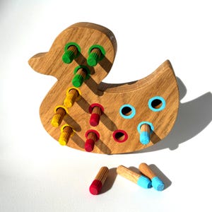 Puede incluir: Juguete de madera con forma de pato y clavijas de colores. El pato es de madera marrón claro y tiene agujeros con clavijas verdes, amarillas y rojas. Clavijas adicionales con puntas azules y rojas están dispersas cerca. Un juguete divertido y educativo.