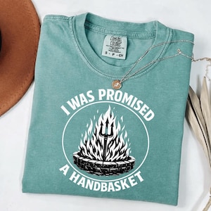 May include: A teal Comfort Colors t-shirt with the text "I WAS PROMISED A HANDBASKET" in a white circular design. The design features a trident in flames. A silver necklace with a pendant is also visible.