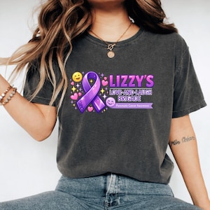 May include: Dark gray t-shirt featuring the text "Lizzy's Love-and-Laugh Brigade" with a purple ribbon and the words "Pancreatic Cancer Awareness." The design includes hearts, stars, and smiley faces.