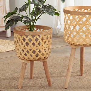 Op de afbeelding: Twee geweven bamboe plantenstandaards met houten poten. Eén standaard bevat een groene plant met bladeren. De standaards hebben een natuurlijke houten afwerking en een ruitpatroon. De standaards staan op een neutraal gekleurd tapijt.