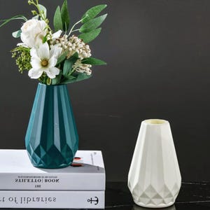 Peut inclure: Deux vases décoratifs. L'un est de couleur teal et contient des fleurs blanches et de la verdure. L'autre est blanc et vide. Les deux vases ont un design géométrique. Les vases sont posés sur deux livres.