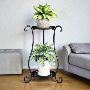 Op de afbeelding: Een zwarte plantenstandaard met twee niveaus en decoratieve krullen. De standaard heeft twee potplanten: een met lichtgroene bladeren en de andere met groen-wit gestreepte bladeren. De standaard is van metaal.