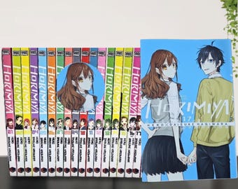 Puede incluir: Una colección de volúmenes de manga "Horimiya". Los lomos muestran el título en blanco y los números de volumen. La portada del volumen 17 presenta a una pareja tomados de la mano sobre un fondo azul. También se ve una pegatina redonda con un retrato de un personaje.
