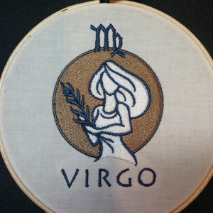 May include: Embroidered Virgo zodiac sign on a white fabric with a brown wooden hoop. The sign features a stylized woman holding a branch with leaves, surrounded by a brown circle. The word "VIRGO" is written in blue below the image.
