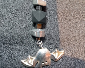 May include: A silver-colored bat-shaped keychain charm with a ring at the top. The charm is connected to a series of black and silver beads. The keychain is designed with a Halloween theme.
