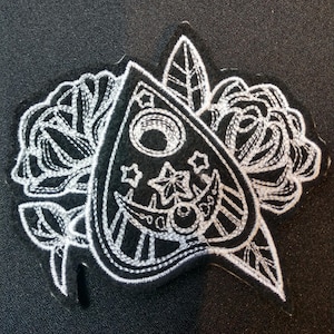 May include: A black and white embroidered patch featuring a white outline of a  witch's  pendulum with a crescent moon and stars. The pendulum is surrounded by two white outlined roses.