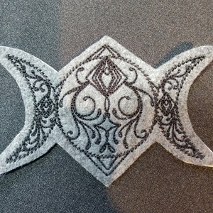 May include: A black embroidered triple moon symbol patch on a gray felt background. The symbol is a stylized representation of the moon in its three phases: new moon, crescent moon, and full moon.