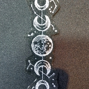May include: A black felt patch with white embroidery of five crescent moons and a circle with a floral design. The moons are connected by lines and dots.
