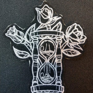 May include: A black and white embroidered patch featuring an hourglass with three roses around it. The roses are stylized and have a vintage look.
