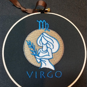 May include: Embroidered Virgo zodiac sign on a black fabric background with a brown wooden hoop. The sign features a stylized image of a woman holding a branch with leaves, with the word "VIRGO" embroidered below.