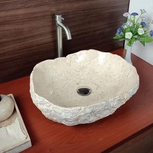 Puede incluir: Un lavabo de piedra beige de forma ovalada con una textura exterior rugosa y natural y un interior liso. El lavabo cuenta con un desagüe central cromado y se combina con un grifo alto y delgado de metal cepillado montado detrás.