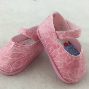 May include: A pair of pink canvas shoes with a floral pattern. The shoes have a strap that goes around the ankle and a buckle closure.