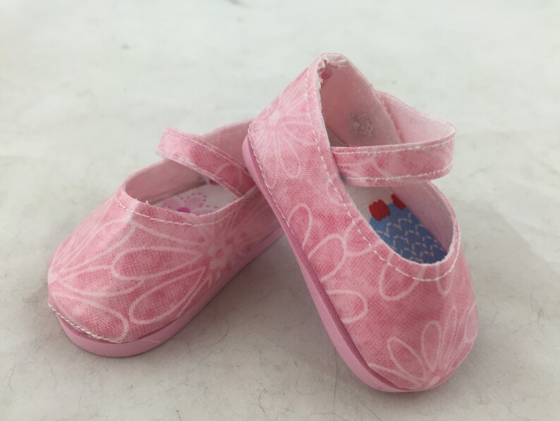 May include: A pair of pink canvas shoes with a floral pattern. The shoes have a strap that goes around the ankle and a buckle closure.
