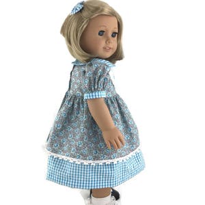 K&ouml;nnte beinhalten: Eine Puppe tr&auml;gt ein blaues und wei&szlig;es Gingham-Kleid mit einem floralen Oberteil und wei&szlig;er Spitzenborte. Die Puppe hat blonde Haare und blaue Augen und tr&auml;gt wei&szlig;e Socken und schwarze Schuhe.