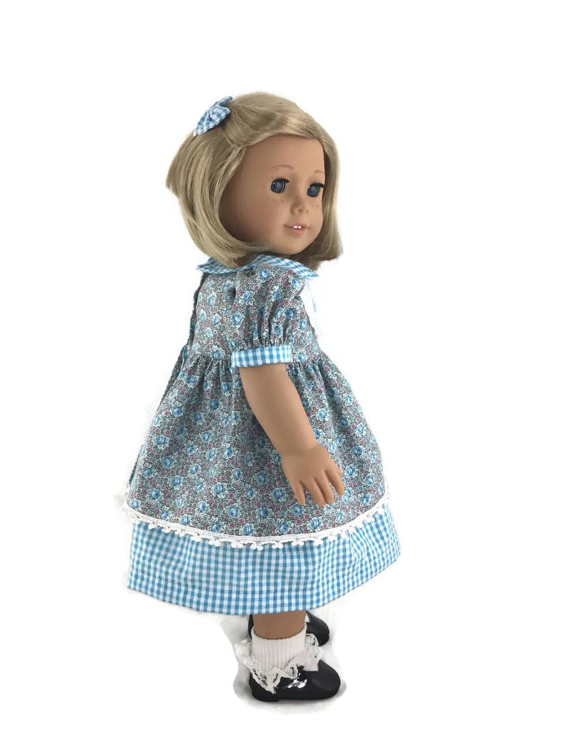 K&ouml;nnte beinhalten: Eine Puppe tr&auml;gt ein blaues und wei&szlig;es Gingham-Kleid mit einem floralen Oberteil und wei&szlig;er Spitzenborte. Die Puppe hat blonde Haare und blaue Augen und tr&auml;gt wei&szlig;e Socken und schwarze Schuhe.