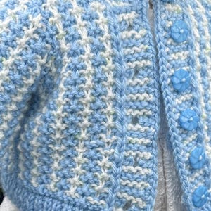 May include: A light blue and white knitted cardigan with a textured pattern. The cardigan features a round neckline, long sleeves, and is fastened with three light blue flower-shaped buttons. The garment is designed for a child.