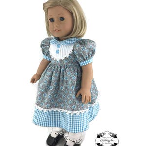 K&ouml;nnte beinhalten: Eine Puppe tr&auml;gt ein blaues und wei&szlig;es Gingham-Kleid mit einem floralen Oberteil und wei&szlig;er Spitzenverzierung. Das Kleid hat Puff&auml;rmel und einen wei&szlig;en Kragen mit blauem Besatz. Die Puppe tr&auml;gt wei&szlig;e Socken und schwarze Schuhe.