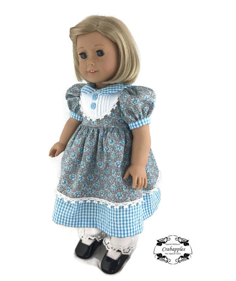 K&ouml;nnte beinhalten: Eine Puppe tr&auml;gt ein blaues und wei&szlig;es Gingham-Kleid mit einem floralen Oberteil und wei&szlig;er Spitzenverzierung. Das Kleid hat Puff&auml;rmel und einen wei&szlig;en Kragen mit blauem Besatz. Die Puppe tr&auml;gt wei&szlig;e Socken und schwarze Schuhe.