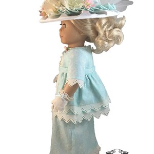 May include: A doll wearing a light blue dress with white lace trim and a white hat with floral embellishments. The doll has blonde hair and blue eyes.