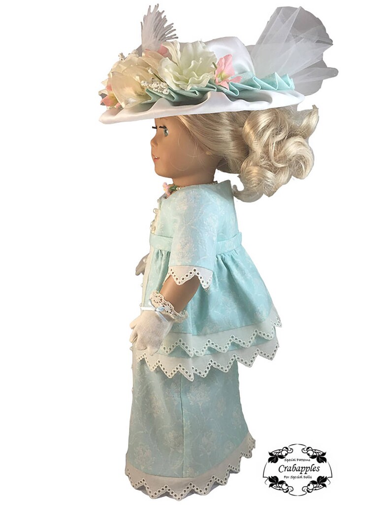 May include: A doll wearing a light blue dress with white lace trim and a white hat with floral embellishments. The doll has blonde hair and blue eyes.