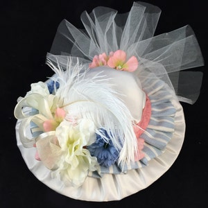 May include: A white hat with a pink and blue striped ribbon, white feather, and white tulle. The hat is decorated with white, pink, and blue flowers.