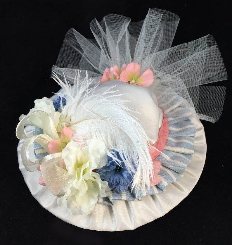 May include: A white hat with a pink and blue striped ribbon, white feather, and white tulle. The hat is decorated with white, pink, and blue flowers.
