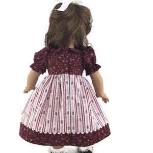 K&ouml;nnte beinhalten: Ein burgunderfarbenes und wei&szlig;es Blumenkleid mit wei&szlig;er Spitzenborte. Das Kleid hat ein tailliertes Oberteil und einen vollen Rock. Die Puppe tr&auml;gt wei&szlig;e Socken und schwarze Schuhe.
