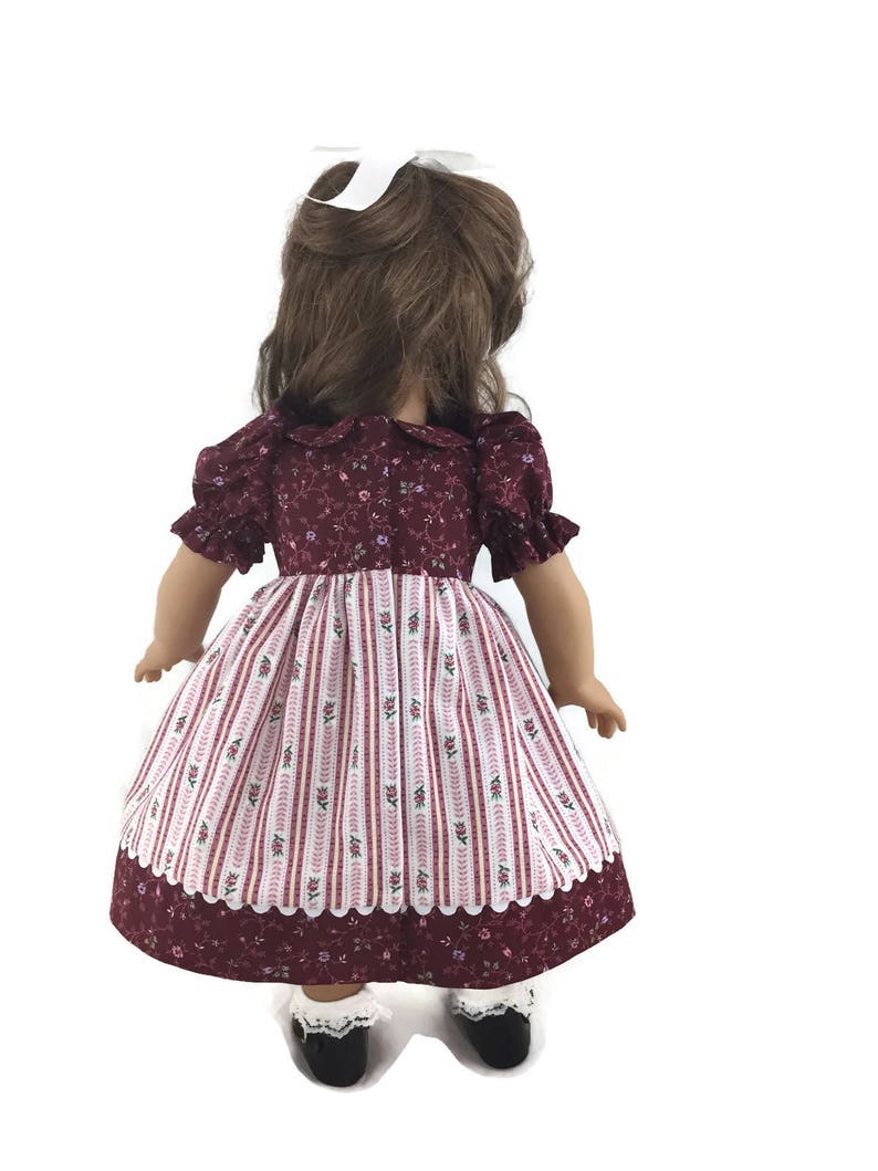 K&ouml;nnte beinhalten: Ein burgunderfarbenes und wei&szlig;es Blumenkleid mit wei&szlig;er Spitzenborte. Das Kleid hat ein tailliertes Oberteil und einen vollen Rock. Die Puppe tr&auml;gt wei&szlig;e Socken und schwarze Schuhe.