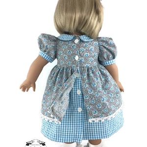 K&ouml;nnte beinhalten: Ein blaues und wei&szlig;es Gingham-Kleid mit einem floralen Oberteil und wei&szlig;em Spitzenbesatz. Das Kleid hat einen Peter-Pan-Kragen und Puff&auml;rmel. Die Puppe tr&auml;gt wei&szlig;e Socken und schwarze Schuhe.