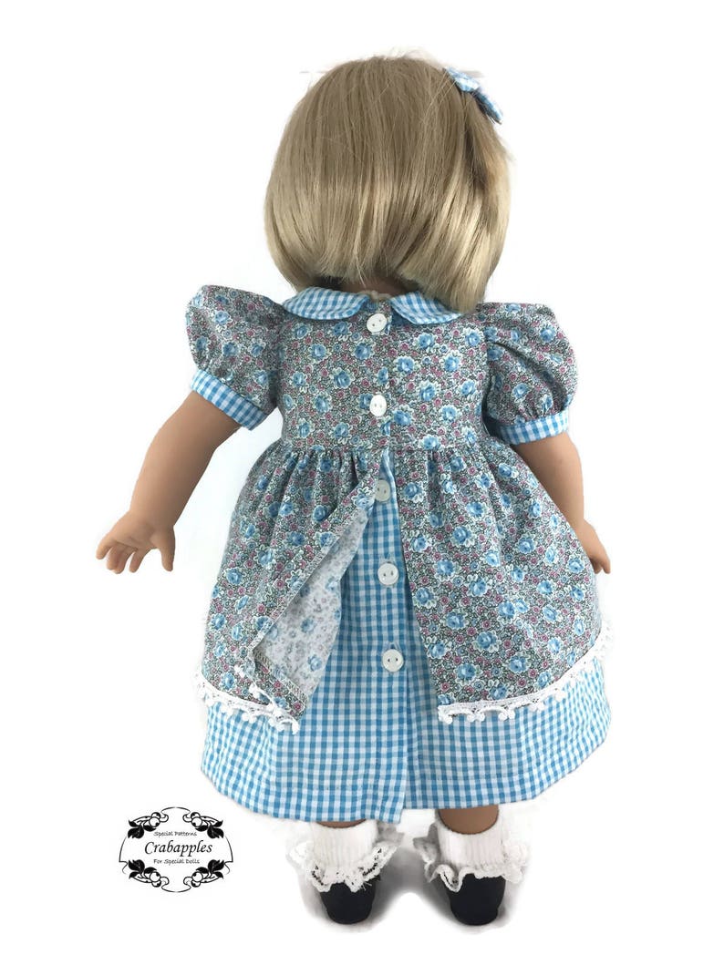 K&ouml;nnte beinhalten: Ein blaues und wei&szlig;es Gingham-Kleid mit einem floralen Oberteil und wei&szlig;em Spitzenbesatz. Das Kleid hat einen Peter-Pan-Kragen und Puff&auml;rmel. Die Puppe tr&auml;gt wei&szlig;e Socken und schwarze Schuhe.