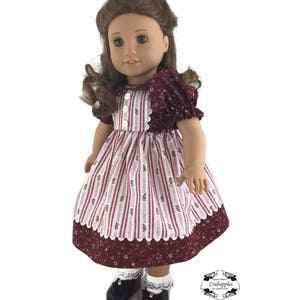K&ouml;nnte beinhalten: Eine Puppe tr&auml;gt ein burgunderfarbenes und wei&szlig; gestreiftes Kleid mit Blumenmuster. Das Kleid hat einen wei&szlig;en Spitzenbesatz und die Puppe tr&auml;gt schwarze Schuhe und wei&szlig;e Socken mit Spitzenbesatz.