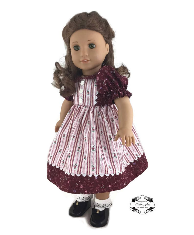 K&ouml;nnte beinhalten: Eine Puppe tr&auml;gt ein burgunderfarbenes und wei&szlig; gestreiftes Kleid mit Blumenmuster. Das Kleid hat einen wei&szlig;en Spitzenbesatz und die Puppe tr&auml;gt schwarze Schuhe und wei&szlig;e Socken mit Spitzenbesatz.