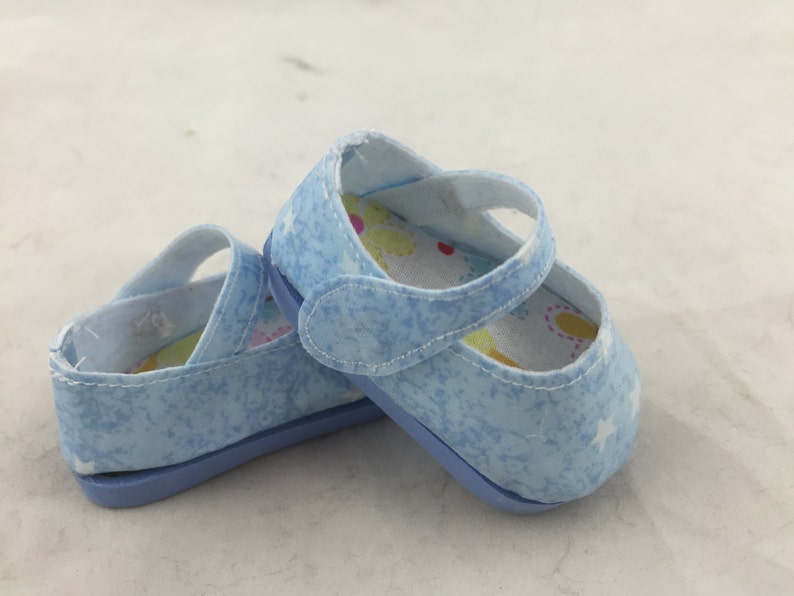 May include: A pair of light blue baby shoes with a white star pattern and a white lining with colorful flowers. The shoes have a velcro strap closure.
